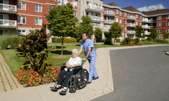 An elderly woman being pushed in a wheelchair by a staff member at a long term care home.