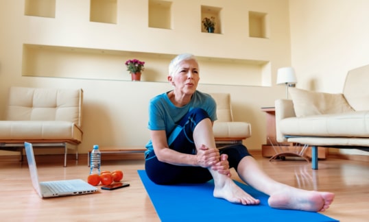Senior woman sitting on the mat, grabbing an ankle, unable to start yoga work out because of sport injury, feeling pain.