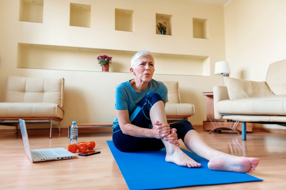 Senior woman sitting on the mat, grabbing an ankle, unable to start yoga work out because of sport injury, feeling pain.