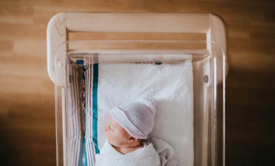 A baby just born at the hospital rests in a hospital bassinet crib, wrapped in a swaddle and wearing a beanie hat.