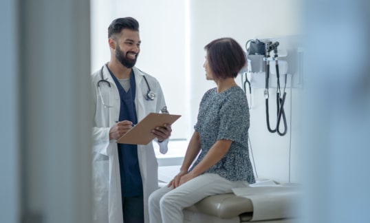 A senior woman sit up on the exam table as she talks with her male doctor about the benefits of the Flu Shot. She is dressed casually and listening attentively as her doctor holds out a clipboard of information and they discuss it.