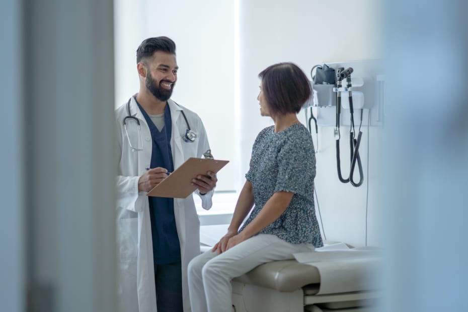 A senior woman sit up on the exam table as she talks with her male doctor about the benefits of the Flu Shot. She is dressed casually and listening attentively as her doctor holds out a clipboard of information and they discuss it.