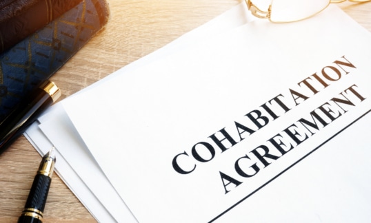 Paper document on a wood surface next to glasses, books, and a black pen. The paper has large black leaders reading Cohabitation Agreement.