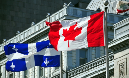 Quebec flag and Canada flag in flying in the wind in front of a building.
