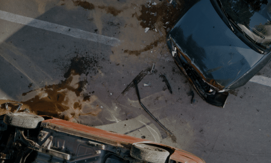 Bird's eye view of two cars in an accident. An orange one is flipped over and the black one has a crushed front. Dirt a and debris sprinkle the ground.