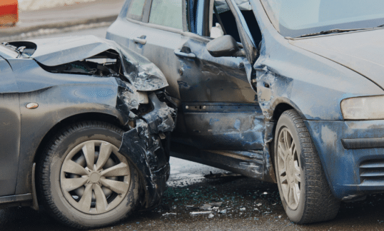 Collision of two blue grey cars denting the hood of one vehicle and the door of another vehicle. Glass shards sprinkled on the pavement below the impact.